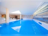 Indendørs swimmingpool med lyseblåt vand, hvide liggestole langs den ene side og en moderne flisebelagt væg. Ovenlysvinduet lukker naturligt lys ind.