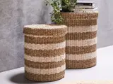 To cylindriske flettede kurve med hvide og naturbrune vandrette striber, den ene brugt som bord med bøger og en potteplante, placeret op ad en grå væg.