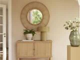 Et træskab med en lampe, en plante og en bog står under et rundt rottingspejl på en beige væg; en stor vase med blomster står til højre.