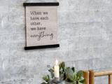 På et skilt på en grå væg står der: Når vi har hinanden, har vi alt. Nedenfor ses et tændt stearinlys og en grøn plante i forgrunden.