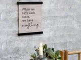 På et skilt på en grå væg står der: Når vi har hinanden, har vi alt. Nedenfor ses et tændt stearinlys og en grøn plante i forgrunden.