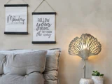 Et hyggeligt hjørne i stuen med en grå polstret sofa, to inspirerende vægtæpper, en dekorativ bladformet væglampe og en lille plante på et bord.