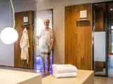 En mand i ternede shorts kommer smilende ud af en dampende sauna. En badekåbe hænger ved siden af døren, og en stak hvide håndklæder ligger på en bænk i et moderne spa-miljø.