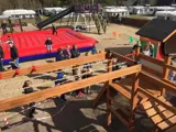 Børn og voksne leger på en legeplads med trækonstruktioner, en stor rød hoppemåtte og rutsjebaner, omgivet af sand og mobilhomes.