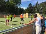 Flere personer spiller fodbold på en udendørs bane iført farvestrålende veste, mens et par tilskuere ser på bag et træhegn under en solrig himmel.