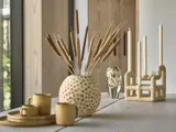 Et træbord med dekorative genstande: en rund vase med tørrede planter, en skulpturel lysestage med tre lys, to gule kopper, træbakker og en glasvase.