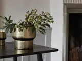 En krukke med guldstruktur og grønne og hvide blomster står på et mørkt bord nær en pejs i et dæmpet oplyst rum.