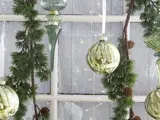 Grønne glasornamenter og fyrretræsguirlande med små kogler hænger foran et matteret vindue og skaber en festlig juledekoration.