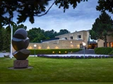 En moderne bygning med beige murstensvægge, omgivet af træer og en pænt trimmet græsplæne med en stor abstrakt skulptur og runde havelamper.