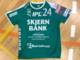En grøn Skjern Bank-håndboldtrøje med hvid tekst, logoer og sponsorer ligger fladt på et trægulv i en gymnastiksal med synlige banelinjer.
