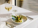 En tallerken med salat står på en hvid dug ved siden af et glas hvidvin, et glas vand, en gaffel og en kniv. Sollyset strømmer ind fra et vindue i nærheden.