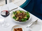 En tallerken med blandet grøn salat med dressing står på en hvid dug ved siden af et glas rødvin, en tallerken med brød og sølvtøj.