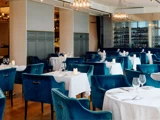 Elegant restaurantinteriør med blå fløjlsstole, hvide duge, dækkede borde, vinglas og blød belysning fra moderne lysekroner.
