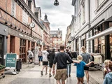 Folk går ned ad en gågade med butikker og caféer på en overskyet dag i en europæisk by med historiske murstensbygninger i baggrunden.