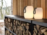 To glødende lanterner med håndtag står oven på sorte hylder fyldt med brænde op ad en trævæg på en udendørs terrasse.