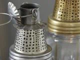 Nærbillede af to vintage lanterner af metal og glas med perforerede toppe; en sølvfarvet i forgrunden og en messingfarvet i baggrunden.