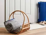 En flettet kurv med hank rummer en sammenrullet blomstret dyne og magasiner, placeret på et trægulv nær en hvid dør og en blå radiator.