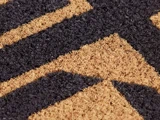 Nærbillede af en dørmåtte med grov kokosstruktur og et sort og brunt geometrisk mønster med diagonale linjer og former.