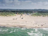 En bred sandstrand med blide bølger, et par både og mennesker spredt langs kysten og en lille kystby med grønne områder i baggrunden under en delvis overskyet himmel.