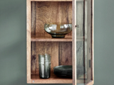 Et vægskab i træ med en glasdør, der viser en glasskål, et glasglas og en stak mørke tallerkener på to hylder.