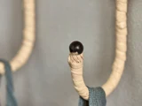 Et nærbillede af en krog, der er viklet ind i beige sejlgarn og har en mørk, rund spids. Et blågråt stykke stof hænger ned fra krogen foran en grå væg.