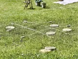 Stort tic-tac-toe-spil på græs med reb som gitter og træskiver som spillebrikker; havemiljø i baggrunden.