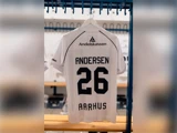 En hvid fodboldtrøje, der hænger i et omklædningsrum, med navnet ANDERSEN og nummer 26 på ryggen.