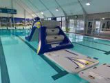 En stor oppustelig rutsjebane og en flydende platform er sat op i en indendørs swimmingpool med højt til loftet og store vinduer.