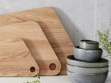 To skærebrætter af træ og en stak med fire grå keramikskåle står på et køkkenbord op ad en betonvæg. Nogle grønne krydderurter står i nærheden.