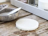 En rund, hvid rensesvamp ligger på en træplade ved siden af en grå skål med en spatel og en hvid bakke med hudplejeprodukter.