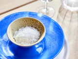En lille skål fyldt med flager af havsalt står på en blå tallerken med et glas og en vandkande i baggrunden.