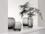 Fire gennemskinnelige grå glasvaser er arrangeret på en hvid hylde, den ene med sarte grønne blade, mod en minimal hvid baggrund.