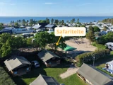 Luftfoto af en campingplads ved havet med telte, legeplads og et skilt, der peger mod et safaritelte blandt træerne tæt på legepladsen.