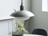 En moderne pendel hænger over et rundt hvidt bord med en sort vase med grønne og hvide blade. En sort stol er delvist synlig i baggrunden.