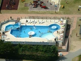 Luftfoto af en swimmingpool med folk, der svømmer og slapper af, omgivet af et solbadningsområde; en legeplads og græs er synlige i nærheden.