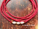 Nærbillede af et wrap-armbånd i fire lag med små røde perler og tre hvide ovale perler, placeret på en rustik brun overflade. Teksten lyder 4 x wrap-armbånd.