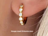 Nærbillede af et øre med en guldring med zirkonia-sten. Teksten nedenunder lyder: Hoops med Zirkonia sten.
