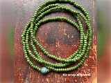 Et grønt perlearmbånd med guldaccenter og en enkelt blå perle, viklet i fire omgange, på en træoverflade. Teksten lyder 4 x wrap-armbånd.