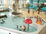 Indendørs swimmingpool med legende børn og voksne, et springvand med røde svampe, vandrutsjebaner og mennesker, der sidder rundt om poolområdet.