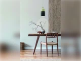 En minimalistisk spiseplads med et træbord, en stol, en vase med nøgne grene, en lille pyntegenstand og en vævet pendel over. Neutrale toner og hyggelig indretning.