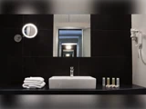 Et moderne badeværelse med en firkantet håndvask, stablede håndklæder, toiletartikler, en væghængt hårtørrer og et rundt, oplyst spejl foran et stort spejl, der reflekterer rummet.