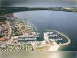 Luftfoto af en kystby med en lystbådehavn, adskillige båd, der ligger fortøjet, bygninger langs kysten og grønne marker i baggrunden.