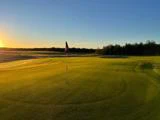 Et golfflag på en putting green ved solopgang, der kaster lange skygger og gyldent lys over det sirligt klippede græs med træer i baggrunden.