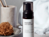 En flaske Meraki Instant Tanning Mousse med sort hætte står på en lys flade ved siden af en naturlig svamp og et hvidt håndklæde i baggrunden.