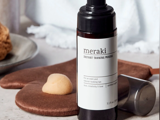 En flaske Meraki Instant Tanning Mousse står ved siden af en solbrændingshandske og en svamp på et badeværelsesbord.