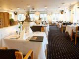 En lys, elegant restaurantspisesal med pænt dækkede borde, hvide duge, træstole og store vinduer, der lukker naturligt lys ind.