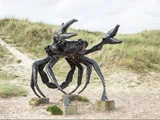 En stor krabbeskulptur i sort metal står på fire betonblokke udendørs med græsklædte sandklitter i baggrunden.