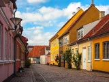En brostensbelagt gade med farverige, gammeldags huse med røde tegltage under en delvis overskyet himmel.