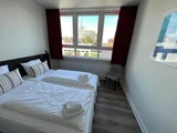 Et pænt hotelværelse med to enkeltsenge, stribede puder, håndklæder, en grå stol, et stort vindue med røde gardiner og en naturskøn udsigt over bygninger og træer udenfor.