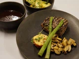 Et tallerkenmåltid med grillet bøf toppet med asparges, ristet tomat, sauterede svampe, en skål brun sauce og en salat med agurk og mikrogrønt ved siden af.